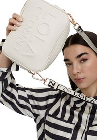 Borsa in eco-pelle bianca con logo impresso, chiusura con zip e tracolla nera removibile con scritte bianche. Blusa a righe con tasche sullo sfondo.