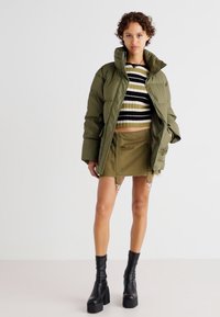 Veste rembourrée vert olive sur un pull cropped rayé noir et blanc et une mini-jupe olive, accompagnée de bottes à talons épais noires.