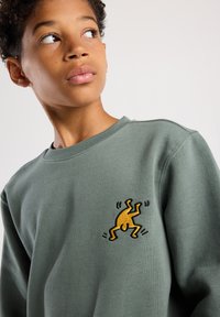 Sudadera verde con una pequeña figura amarilla bordada con contorno negro, cuello redondo, textura suave y corte casual.