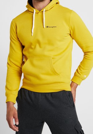 Gelber Pullover-Hoodie mit Fronttasche, weißen Kordeln und schwarzem "Champion"-Logo auf der Brust. Gerippte Bündchen und Saum, weiche Textur.