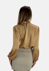 Blusa satinata marrone con maniche a sbuffo, colletto alto e dettaglio a chiave sul retro; abbinata a una gonna aderente beige chiaro.