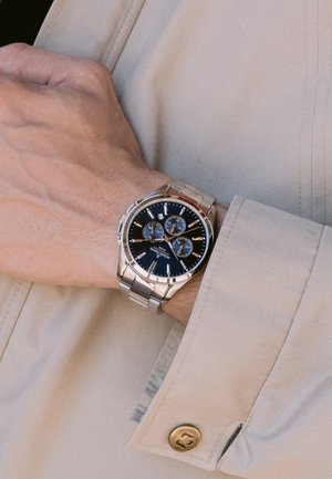 Montre en acier inoxydable argenté avec un cadran noir, trois sous-cadrans chronographes, des accents argentés et un bracelet. Bouton sur le côté pour les réglages.