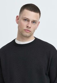 Felpa nera con collo rotondo e tessuto testurizzato. Il modello ha i capelli corti e indossa una camicia bianca visibile al colletto.