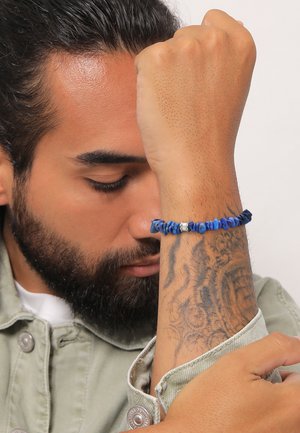 Bracelet en perles bleues aux formes irrégulières, présentant diverses nuances de bleu et des accents argentés, porté sur un avant-bras tatoué.