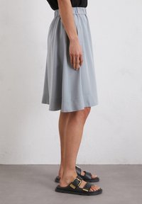 Jupe grise longueur genou avec une taille élastique, dotée de poches latérales et d'une texture lisse. Associée à des sandales foncées avec des accents dorés.