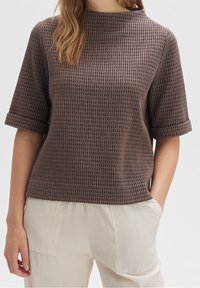 Top corto a maniche corte di colore marrone con texture, collo alto e risvolti, abbinato a pantaloni beige chiari e sgualciti. Vestibilità comoda, design semplice.