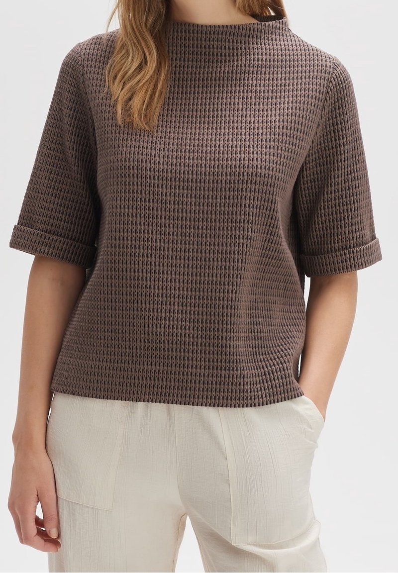Top corto a maniche corte di colore marrone con texture, collo alto e risvolti, abbinato a pantaloni beige chiari e sgualciti. Vestibilità comoda, design semplice.