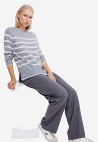 Maglione grigio a strisce con scollatura tonda, abbinato a pantaloni grigi larghi. Il modello è seduto su una panchina bianca, indossando sneaker bianche con un accento a forma di stella.