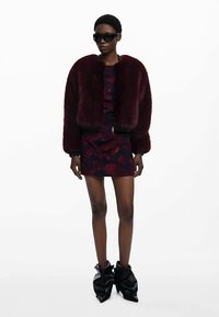 Viininpunainen tekoturkistakki kuosillisen minimekon päällä, yhdistettynä ylisuurin mustiin aurinkolaseihin ja kiiltäviin mustiin nilkkureihin.