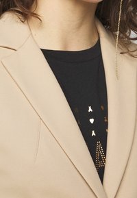 Primer plano de una persona que lleva un blazer beige sobre una camiseta negra con diseños de corazones y estrellas doradas, y un arete de cadena dorada.