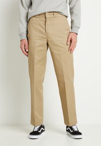 Beige chino kalhoty vyrobené z hladkého materiálu, s přímým střihem a předními záhyby, spárované s černobílými teniskami.
