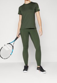 Oliwkowy, sportowy top i legginsy, dopasowany krój, gładka tekstura, w połączeniu z czarnymi trampkami, trzymający rakietę tenisową.