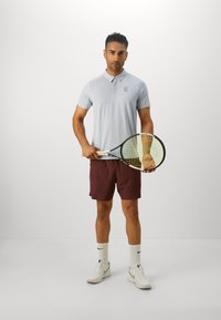 Lyseblå polo med korte ærmer og et logo på brystet, kombineret med maroon shorts. Holder en tennisracket, iført hvide sneakers og sokker.
