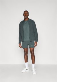 Ciemnozielona polarowa kurtka na zamek, noszona na dopasowanej koszulce i szortach. Białe sneakersy i widoczne skarpetki. Minimalne logo na szortach.