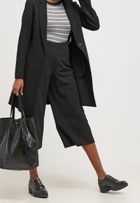 Černý tailorovaný kabát přes vzorovaný top, v kombinaci s širokými černými culottes a černými koženými botami. Součástí je černá kabelka.