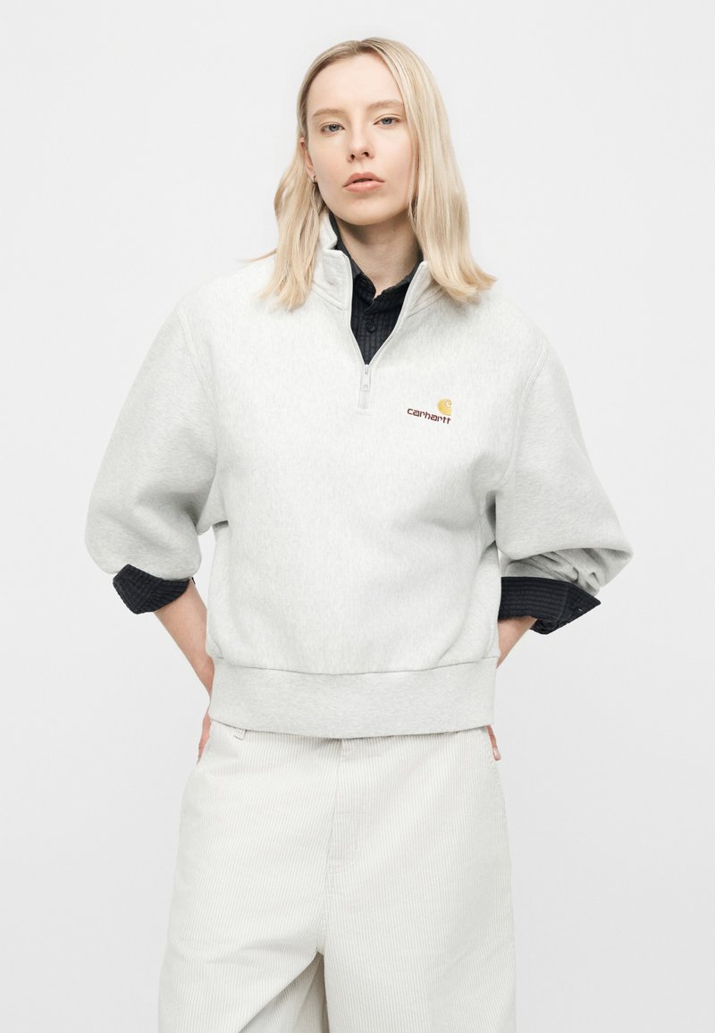 Giovane con capelli biondi che indossa una felpa con zip Carhartt grigio chiaro e pantaloni bianchi, mani posate sui fianchi, sfondo neutro.