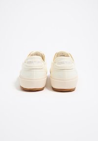 White canvas sneakers with a textured finish, gum rubber soles, and a stitched logo detail on the back. Rounded toe design and low profile.