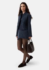 Completo giacca e gonna in tweed texturizzato blu navy, collant neri, mocassini scuri e una borsa intrecciata marrone. Taglio aderente, dettagli minimi.