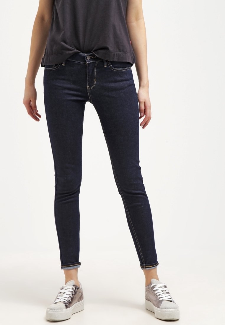 Jean slim bleu foncé, fabriqué en denim, avec un design ajusté, style classique à cinq poches, associé à des baskets argentées.