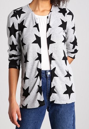 Cardigan grigio con motivo a stelle nere, maniche lunghe e design aperto frontale. Abbinato a una maglietta bianca e jeans blu visibili sotto.