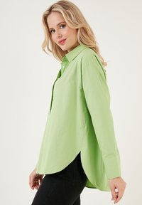 Camicia button-up verde chiaro con una vestibilità rilassata, orlo arrotondato, maniche lunghe e una texture in cotone morbida. Presenta un colletto classico e bottoni frontali.