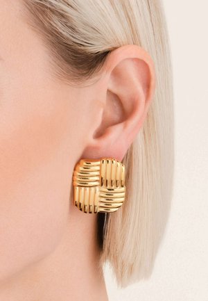 Boucles d'oreilles dorées au design carré, ornées de nervures horizontales et verticales, complétant une oreille lissée et des cheveux blonds courts.