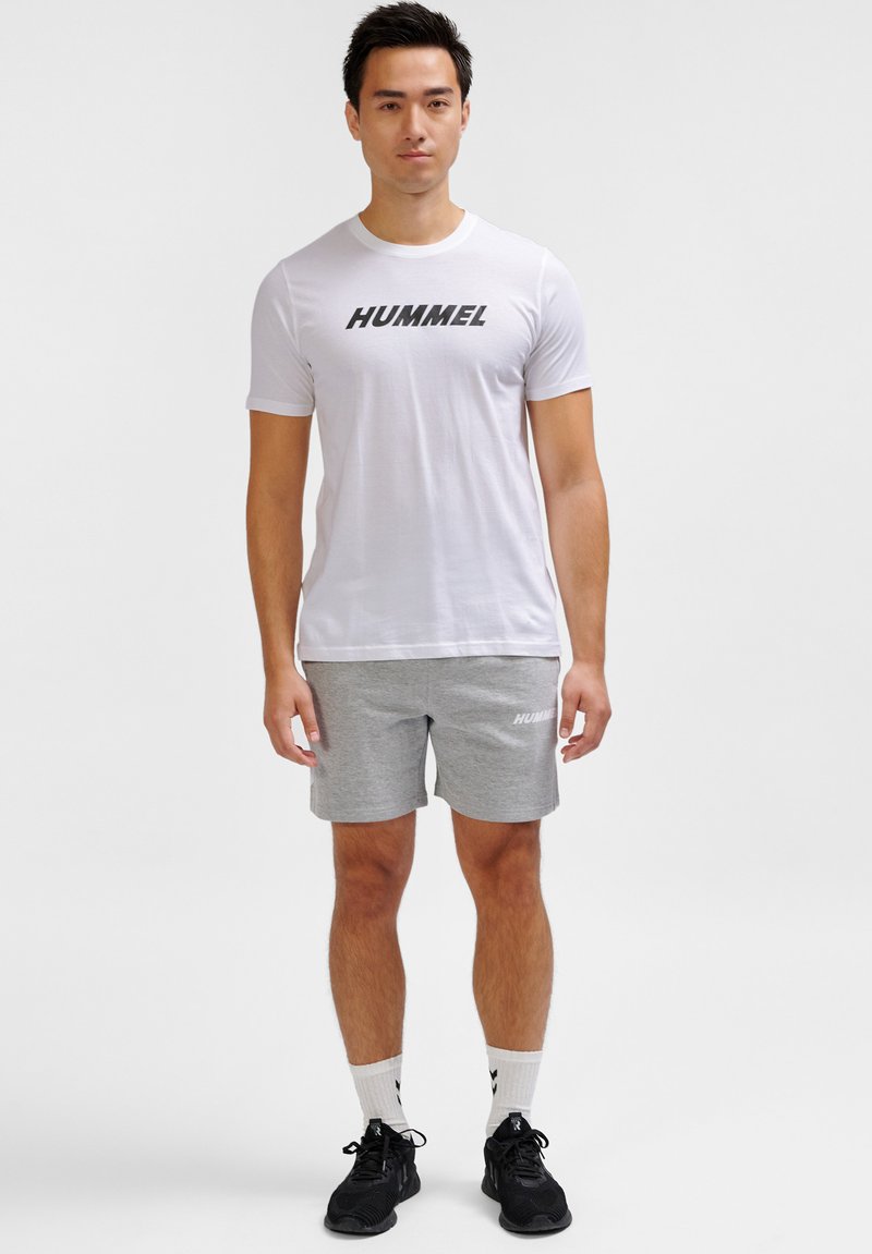 Hvid t-shirt med "HUMMEL" logo, parret med lysegrå shorts. Outfitten har et afslappet design, suppleret af sorte sneakers.