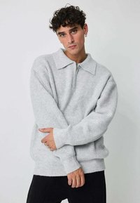 Pull-over tricoté gris clair avec col et demi-fermeture éclair. Manches longues et poignets côtelés. Motif texturé sur l'ensemble.