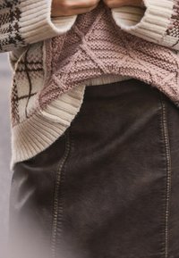 Hænder holder kanten af en lyserød kabelstrikket sweater over en brun læderskirt, med en cremet og brun mønstret cardigan synlig.