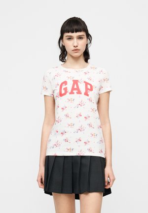 Ung kvinde med kort sort hår iført en hvid blomstret GAP T-shirt og et sort plisseret skørt, stående mod en ensfarvet baggrund.