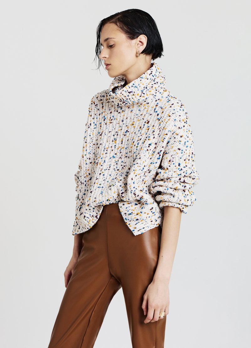 Pullover lavorato a maglia in crema con macchie multicolori, caratterizzato da un collo alto, una vestibilità rilassata e una texture a costine, abbinato a pantaloni di pelle marrone.