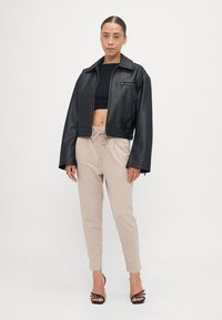 Veste cropped en simili cuir noir avec fermeture éclair et poche poitrine, assortie à un pantalon beige à cordon de serrage et des sandales à talons noires.