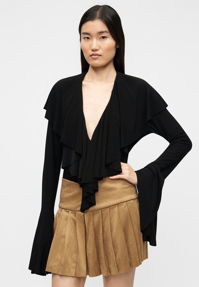 Μαύρο μακρυμάνικο τοπ με βολάν στο γιακά και στη λωρίδα κουμπιών, συνδυασμένο με μίνι φούστα από ταμπά σουέντ με πιέτες. Ομαλή υφή και σχεδιασμός με στρώσεις.