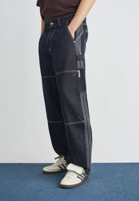 Dunkelblaue Jeans mit weitem Bein, weißen Steppnähten und seitlichen Streifen. Verfügt über seitliche Taschen und eine lockere Passform. Kombiniert mit cremefarbenen Sneakern.