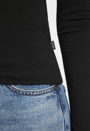 Maglietta nera a maniche lunghe con design aderente, caratterizzata da un piccolo etichetta "GUESS" all'orlo, abbinata a jeans in denim azzurro strappati.
