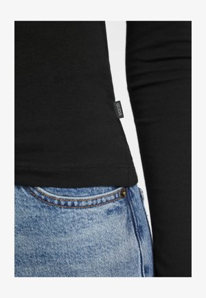 Cămașă neagră cu mâneci lungi, cu un design ajustat, având un mic eticheta "GUESS" la tiv, asortată cu blugi din denim albastru, rupți.