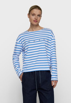 Mujer que lleva una camisa de manga larga a rayas azul y blanca con la palabra "TODAYS" y pantalones azul oscuro, de pie con una mano en el bolsillo.
