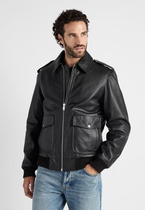 Man met krullend haar die een zwart leren jack met voorzakken en rits draagt, en blauwe jeans, kijkt opzij.