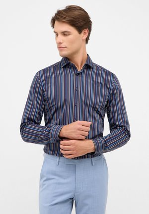 gestreepte shirt in marineblauw, rood en blauw met een button-down kraag en lange mouwen. Draagt met lichtblauwe broek. Glad stoftextuur.