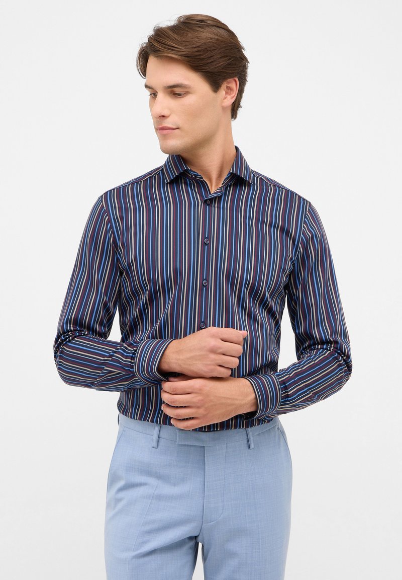 camicia a righe in navy, rosso e blu con colletto button-down e maniche lunghe. Indossata con pantaloni azzurri. Tessuto di superficie liscia.