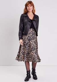 Veste de biker en cuir noir sur un haut noir, associée à une jupe en tan arrivant aux genoux avec un motif léopard noir. Bottes noires à lacets.