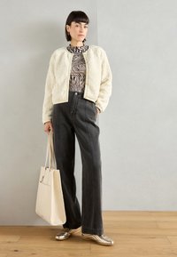 Ivory teddy jacket, black and white patterned top, wide-leg gray jeans, and gold sneakers. Model holds a beige tote bag. Smooth textures.