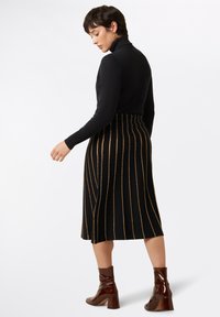 Black turtleneck paired with a knee-length black skirt featuring vertical gold stripes. Model wears glossy brown heeled ankle boots.