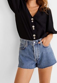 Černá blůza s výstřihem do V a žebrovanou texturou, doplněná pěti světle barevnými knoflíky, spojená s modrými džínovými šortkami s vysokým pasem, které mají ořezaný lem a kapsy.