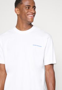 Witte katoen t-shirt met een ronde halslijn, korte mouwen en een kleine blauwe tekstdetails met de tekst "QUEENS BOROUGH" aan de linkerkant.