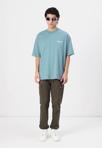 T-shirt oversize bleu clair avec un logo, associé à un pantalon cargo vert olive comportant plusieurs poches, et des baskets blanches.