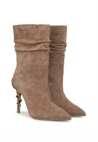 Stivaletti alla caviglia in suede beige con design svasato, punta affusolata e un accento metallico decorativo sul tacco alto. Texture liscia in tutta la superficie.