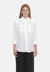 Camicia bianca con colletto, due tasche sul petto con dettagli dorati e maniche lunghe arrotolate ai polsi. Tessuto liscio e leggero.