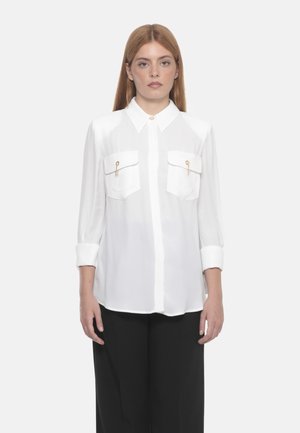 Camicia bianca con colletto, due tasche sul petto con dettagli dorati e maniche lunghe arrotolate ai polsi. Tessuto liscio e leggero.