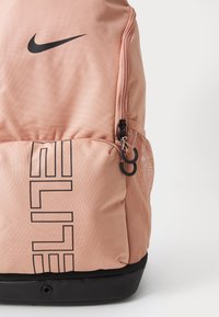 Mochila Nike ELITE en material suave de color rosa claro con acentos negros, que cuenta con un bolsillo lateral de malla, dos compartimentos con cremallera y un detalle del logo.